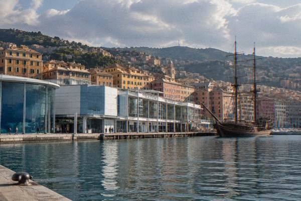 Visitors tiny and distant on a quay outside a long modern waterfront building of white and blue-grey panels, an old wooden sailing ship moored nearby, Aquarium of Genoa, Genoa