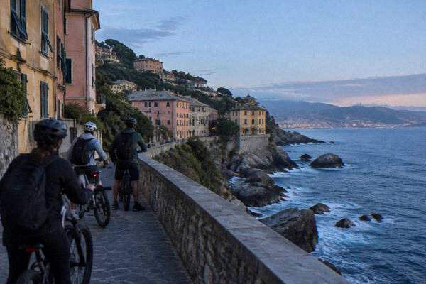 A small group on e-bikes with helmets stopped on a stone-paved cliff-edge promenade above a clear blue sea, Passeggiata Anita Garibaldi