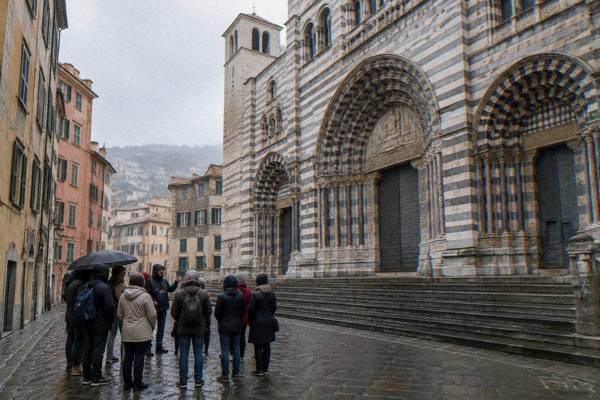 A small walking group gathered round a guide on a narrow sloping square before a cathedral front of striped pale and dark stone, Genoa Cathedral