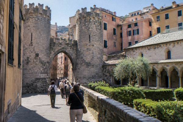 A small walking group passing through a medieval gate of two round stone towers behind a guide, Porta Soprana, Genoa