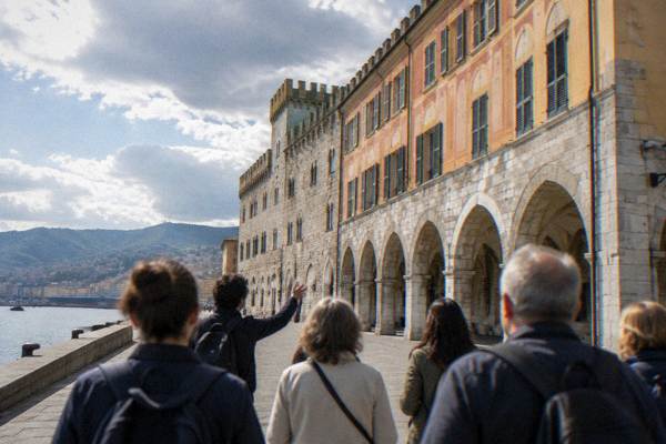 A guide and a small group seen from behind before a long medieval palace front of pale stone and faded painted ochre beside a harbour road, Palazzo San Giorgio