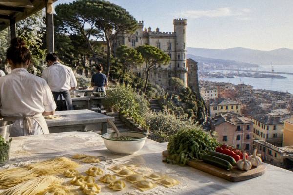 A bright kitchen worktop with fresh pasta shapes, a bowl of green sauce and a wooden board of vegetables, a few cooks seen from behind, Castello D'Albertis