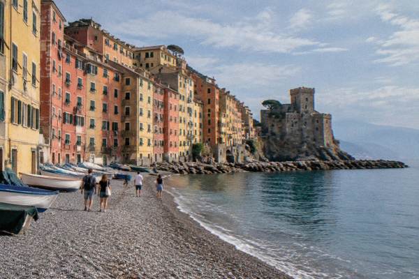 A small group of day visitors walking a pebble beach below very tall slender ochre, red and yellow houses with fishing boats pulled up, Camogli