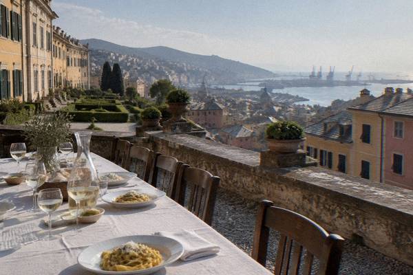 A long table laid for lunch on a terrace with a carafe of white wine, plates of fresh pasta and a view over rooftops and the harbour, nobody seated, Villa del Principe