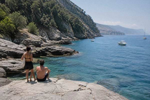 A pair of visitors seen from behind on flat pale rock platforms beside clear blue water below steep wooded cliffs, Punta Chiappa