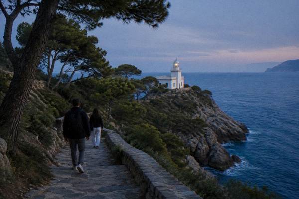 Two visitors seen from behind on a stone path through pines toward a small white lighthouse on a rocky point above deep blue sea, Portofino Lighthouse
