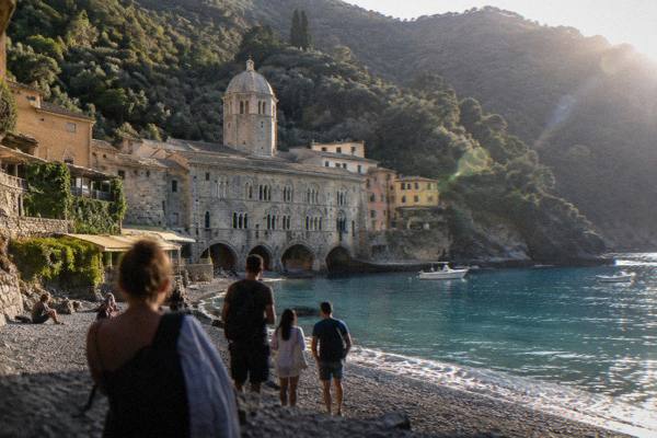 A small group of visitors seen from behind on a shingle beach before a medieval abbey of pale stone in a steep forested cove, San Fruttuoso