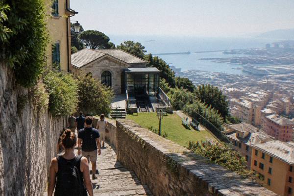 A small group on a hilltop path of old stone paving between high walls, a hazy view of the sea and rooftops far below, Righi Upper Station