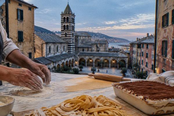 Hands dusting a worktop with flour beside rolled fresh pasta and a tray of tiramisu in a bright kitchen, only hands in the frame, Museo di Sant'Agostino