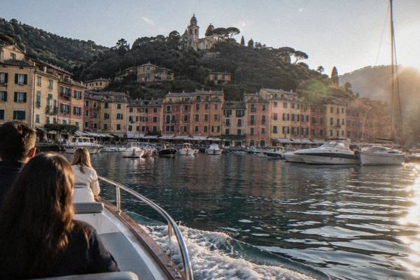 A small motor boat gliding into a tiny horseshoe harbour of pastel houses with a few passengers seen from behind at the rail, Portofino