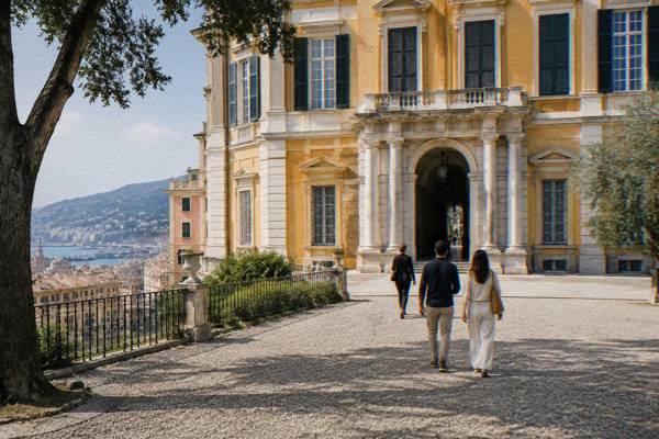 A private guide and a couple seen from behind crossing a gravel forecourt toward a large ochre and white palace, Palazzo Reale