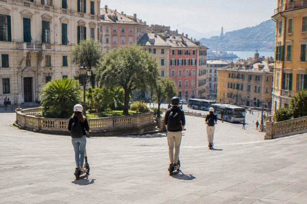 Three riders on two-wheeled standing electric scooters with helmets crossing a wide sloping square of cream stucco palaces, seen from behind, Piazza Corvetto