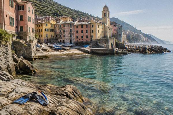 A lone swimmer's snorkel and fins left on warm rocks beside clear shallow sea at a small harbour with pastel houses behind, Vernazzola, Genoa