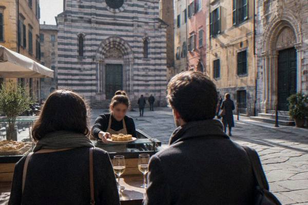 A guide handing a small plate of focaccia across a counter to two visitors seen from behind, glasses of white wine on a high table, Piazza San Matteo