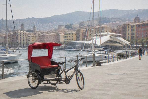 A pedal rickshaw with an empty passenger seat parked on a wide harbour promenade with moored boats and pastel houses behind, Porto Antico