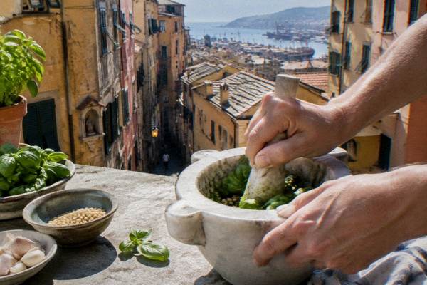 Hands crushing basil, pine nuts and garlic in a marble mortar on a worktop on a sunny rooftop terrace above terracotta roofs, only hands in the frame, Old Town Caruggi