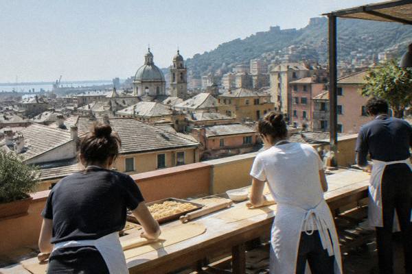 A few people in aprons seen from behind rolling fresh pasta dough on a long worktop on a rooftop terrace with slate rooftops beyond, Genoa City Centre