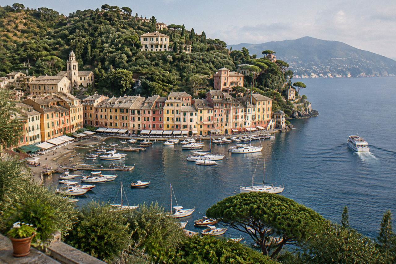 A passenger boat cruising along the Ligurian coast towards a small harbour village backed by wooded hills