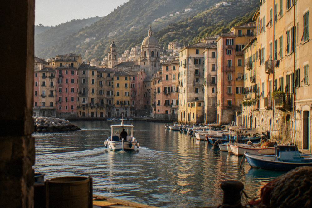 A boat entering a narrow harbour lined with tall pastel buildings and moored fishing dinghies