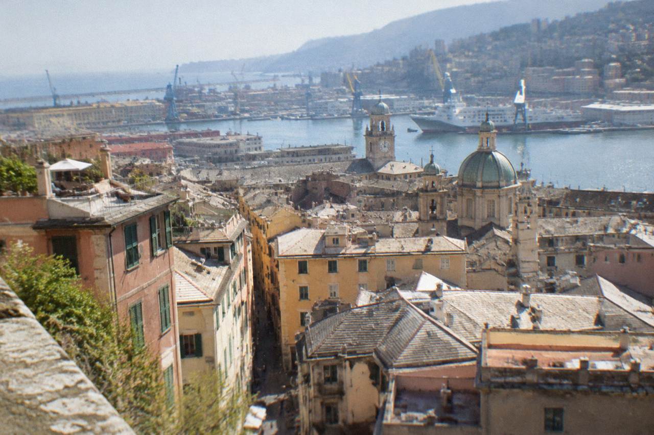 Clock face above rooftops and harbour cranes in Genoa