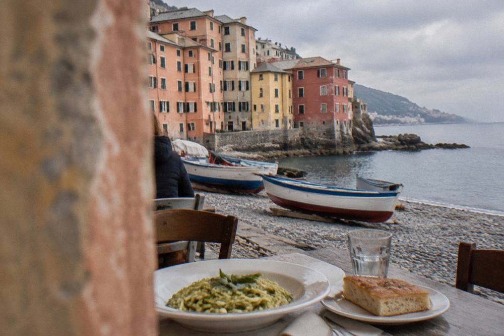 A plate of trofie pasta with green pesto beside a slice of focaccia on a seaside table