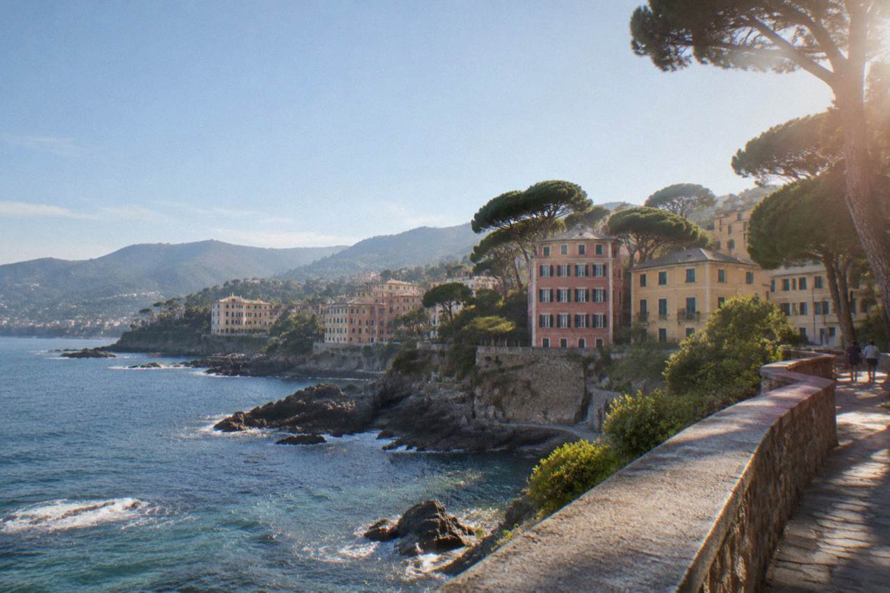 A seaside cliff walk with a low stone railing curving above clear water, pines along the path and hills in the distance