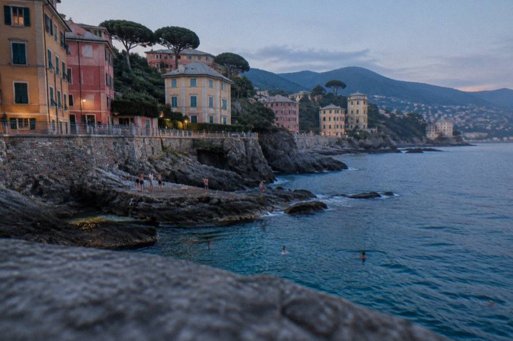 People swimming from flat rocks into clear deep water below a stone promenade wall