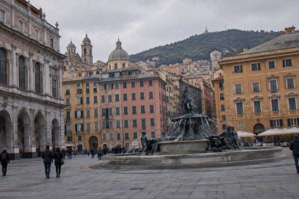 Wide square in central Genoa with a large bronze fountain and historic buildings around it