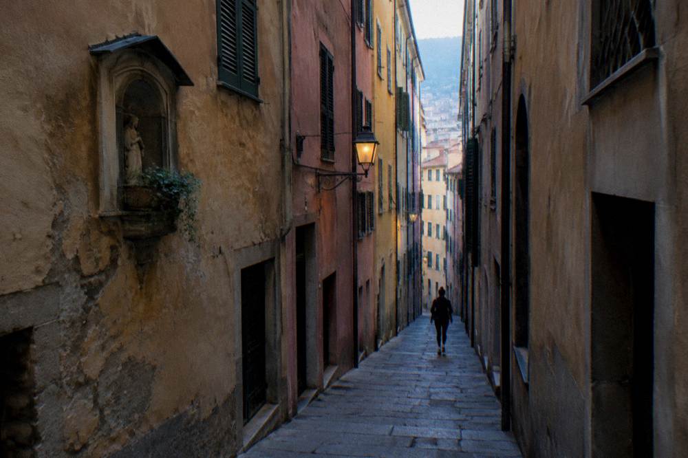 A very narrow deep alley between tall medieval buildings in faded ochre, rose and grey stucco, a worn slate-paved lane and a sliver of sky overhead