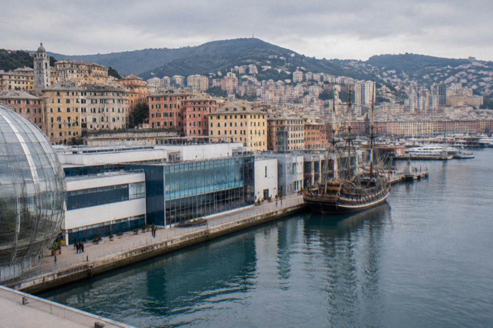 A long low modern waterfront building of white and blue-grey panels and glass beside a quay, calm harbour water and a moored old wooden sailing ship