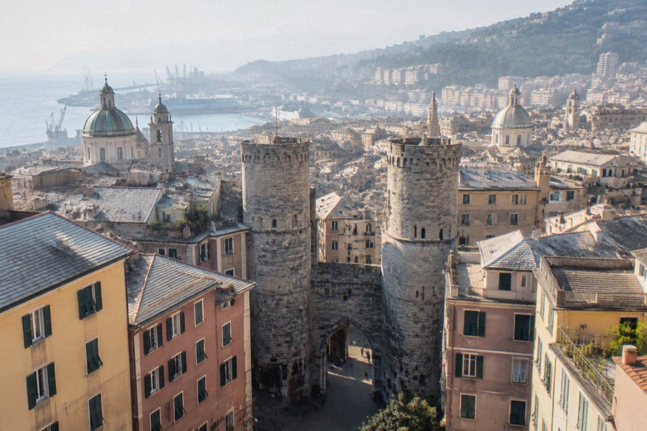 A medieval stone gate with two tall round towers standing above narrow old streets in Genoa