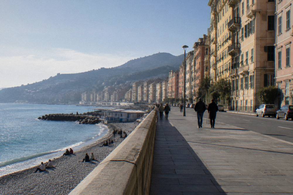 A pebble beach below the promenade with clear water and people sitting on the rocks near bathing establishments