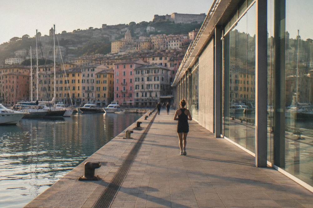 A long modern museum building of glass, pale stone and steel on a dock edge with calm harbour water beside it and pastel houses on the hill behind