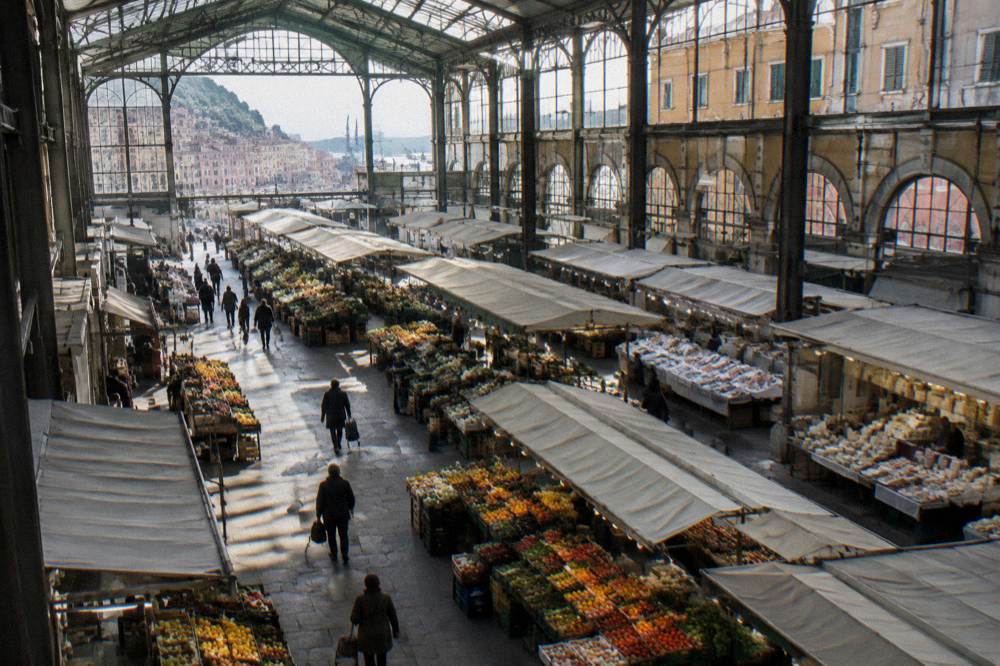 A large covered food market hall with a high iron and glass roof and rows of stalls piled with fruit, vegetables, fish and cheese under plain awnings