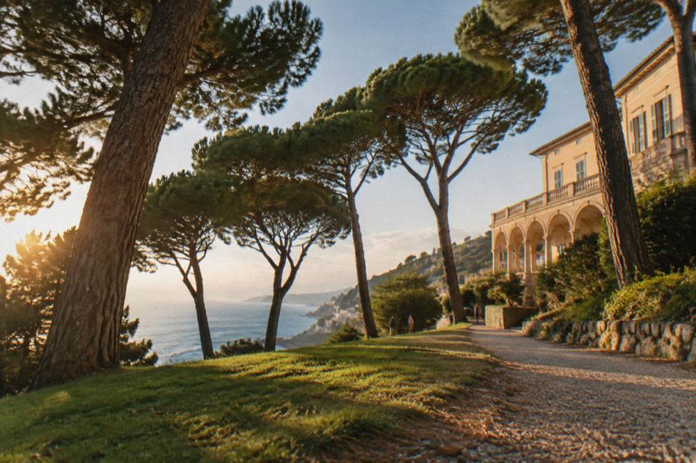 A large landscaped seaside park of lawns, tall umbrella pines and gravel paths with a pale villa and the blue sea visible between the trees