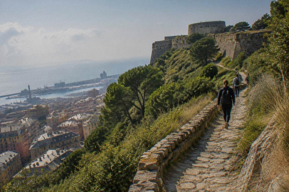 Hikers on a stone path through Mediterranean woodland toward a fort wall