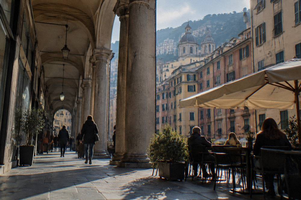 The covered arcade of a Genoa shopping street with columns, shop windows and people seated at a café terrace