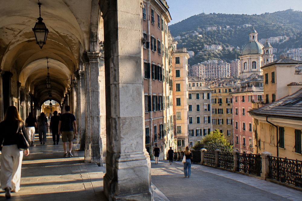 People walking under the stone arcades of a shopping street in Genoa in late afternoon light