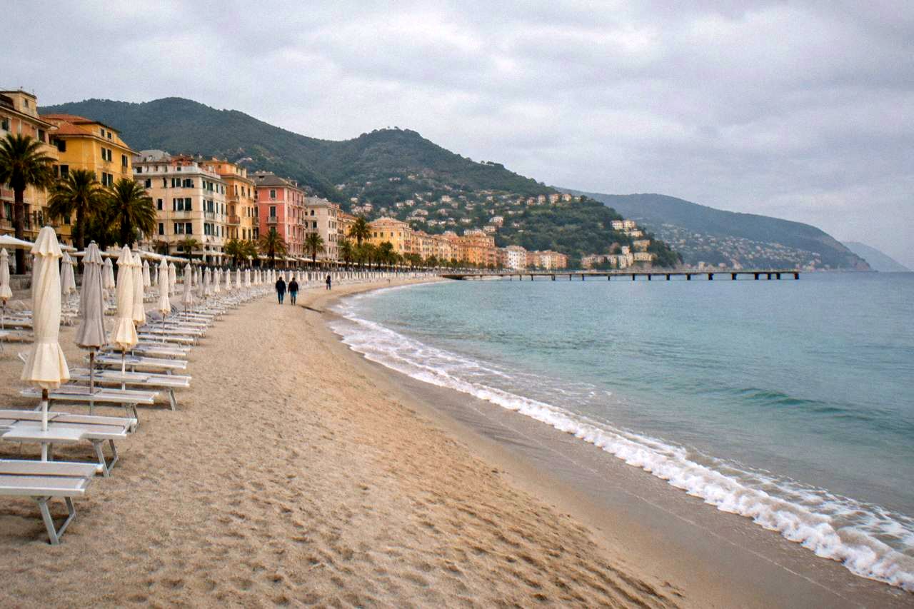 A wide sandy beach curving along a Ligurian seaside town with sun umbrellas and calm blue water