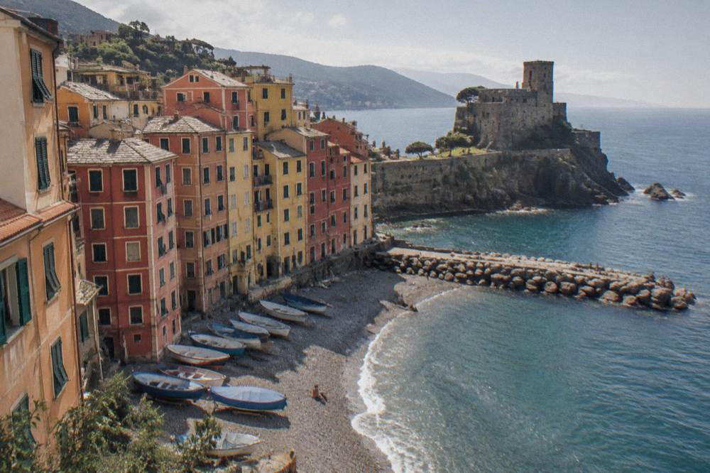 A fishing village of very tall slender houses in ochre, red and yellow stacked above a pebble beach with fishing boats pulled up on the stones