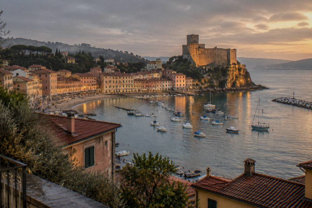 A village harbour at golden hour with castle walls lit above the water