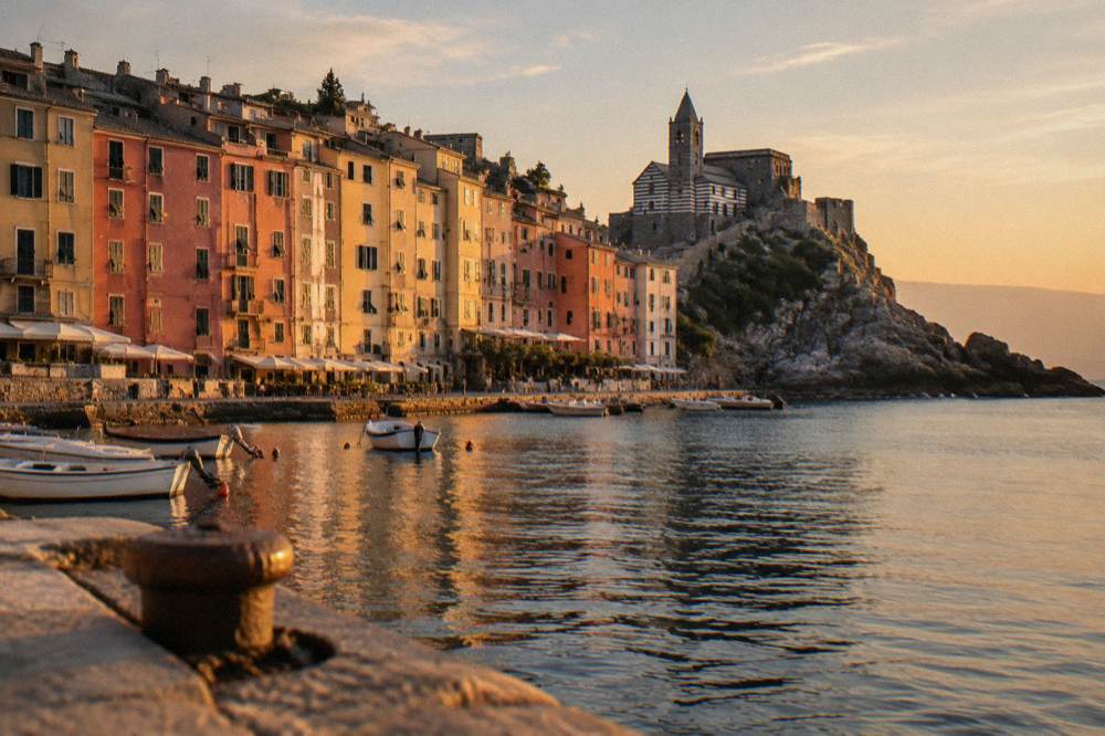 A tall wall of narrow pastel houses along a small harbour with a church of black and white striped stone on a rocky headland at the point