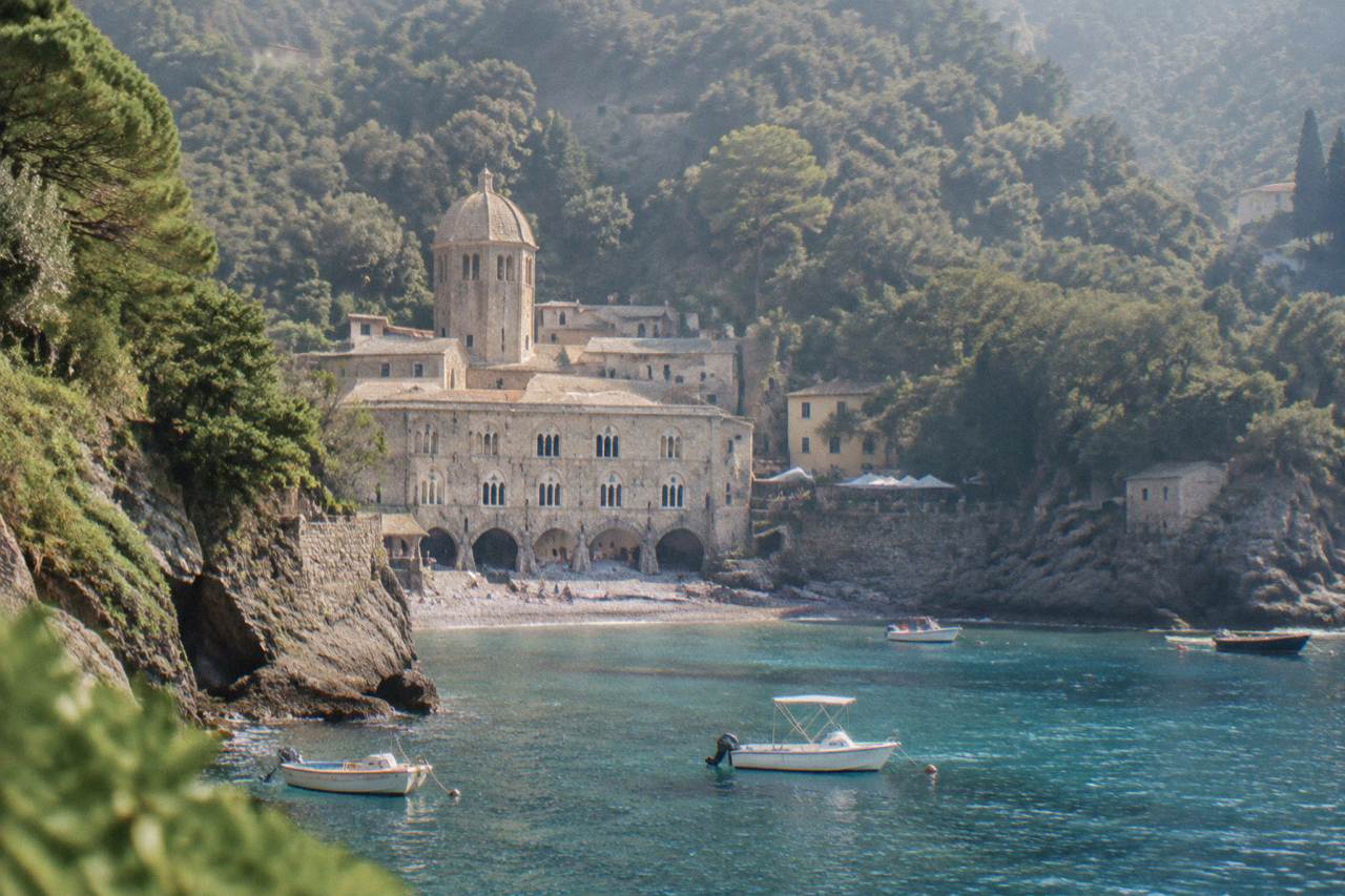 A stone abbey church standing at the head of a small pebble cove with wooded hills rising behind it and small boats moored in the water