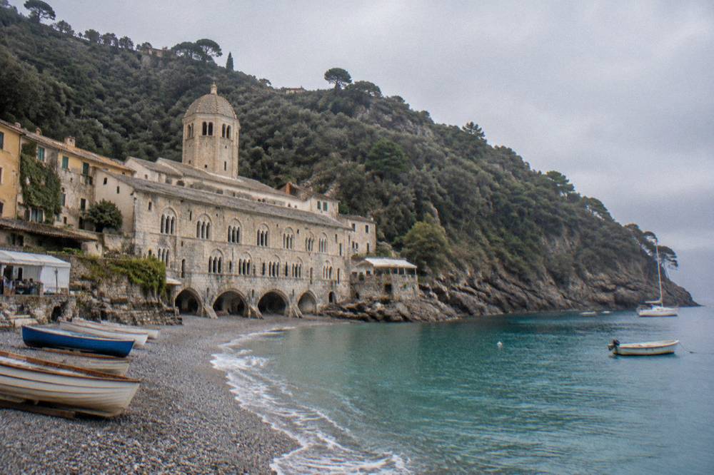 Wooden dinghies pulled up on the pebble beach in front of an abbey facade at midday