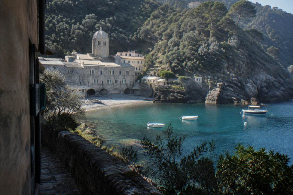 A tiny cove with a medieval abbey of pale stone beside a small shingle beach and steep forested slopes dropping to turquoise water