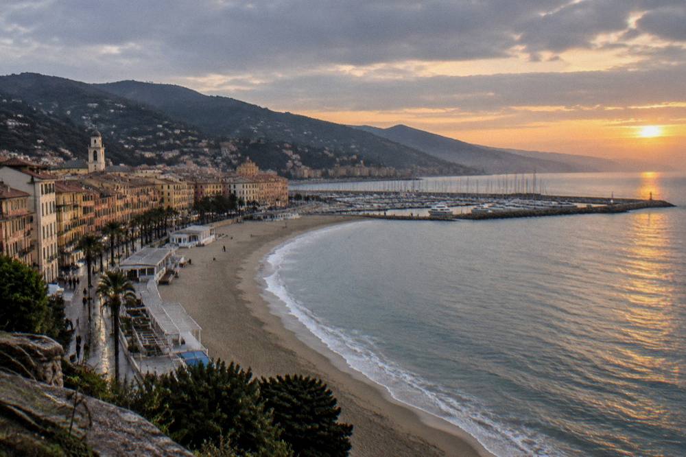 A wide view of the Ligurian coastline with a beach town and hills at sunset