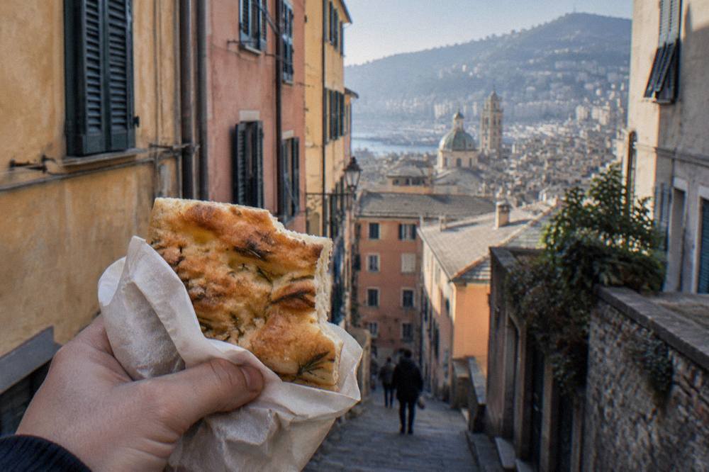 A paperwrapped piece of focaccia held in hand above a narrow old town lane with tall colourful buildings
