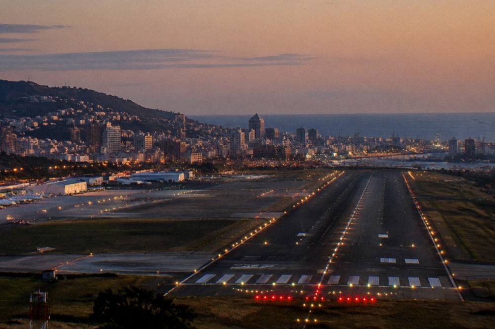 An evening view of an airport runway with the lights of Genoa and the sea in the distance