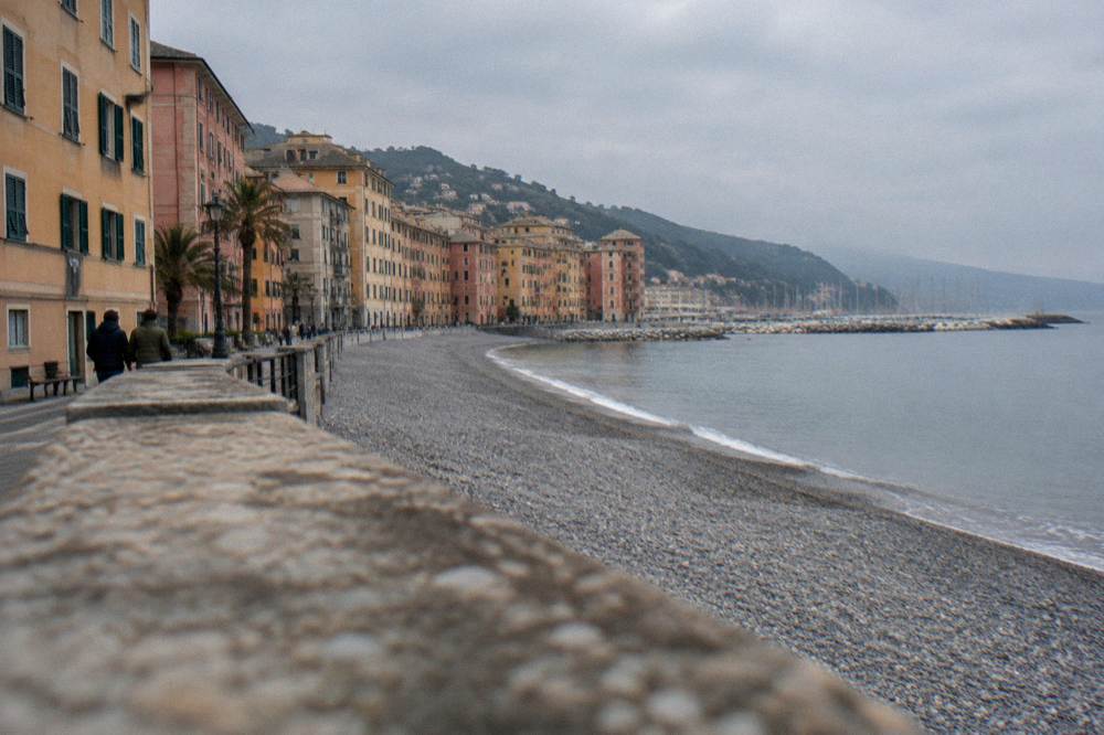 A long pebble beach with calm sea, a promenade of tall pastel buildings and a small marina in the distance