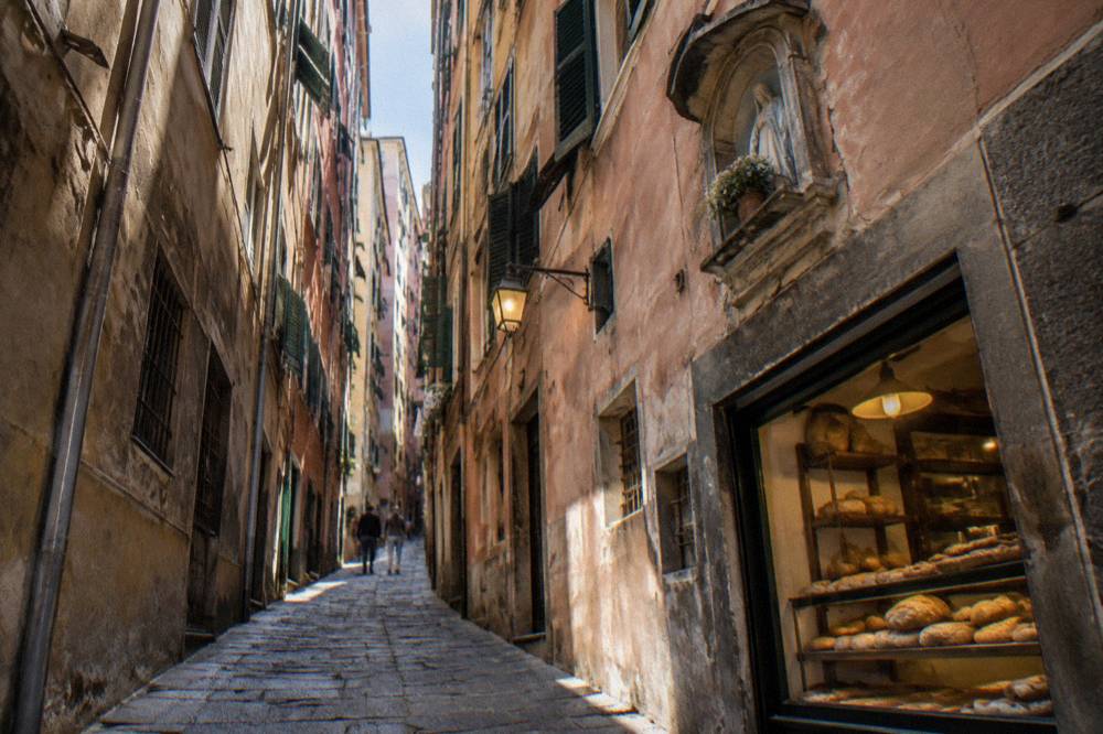 A narrow caruggi lane in Genoa's old town with a small bakery window lit in the morning